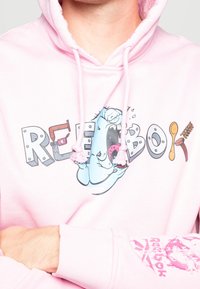 Ljusrosa huvtröja med en stor lekfull grafik av en blå varelse och texten "REEBOK." Har dragsko i huven och ribbade ärmslut.