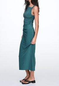 Robe longue sans manches couleur sarcelle avec des fronces à la taille ; tissu lisse et extensible ; associée à des sandales plates noires.