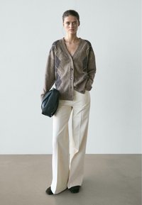 Kobieta stojąca, ubrana w brązowy kardigan w wzory, kremowe spodnie o szerokich nogawkach, czarne buty, trzyma czarną torbę, na jednolitym jasnoszarym tle.