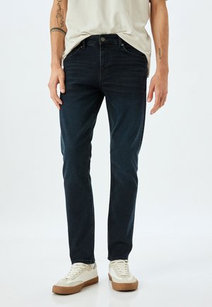 Jeans de denim azul oscuro con un corte ajustado y diseño de cinco bolsillos, combinados con zapatillas crema con una suela de goma marrón.