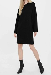 Svart oversized klänning med långa ärmar, tillverkad av ett texturerat tyg, med sidofickor, bärs med spetsiga svarta anklarstövlar.