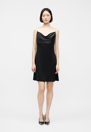 Femme debout en robe noire sans manches avec bretelles fines et décolleté drapé, portant des talons hauts noirs sur un fond uni.