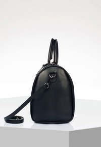 Sac à main en cuir noir avec dessus arrondi, deux poignées et bandoulière amovible, posé sur une surface blanche devant un fond uni.