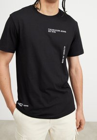 Svart bomullströja med rund hals, med vittryckt text "CALVIN KLEIN JEANS EST 1978" och koordinater på framsidan.