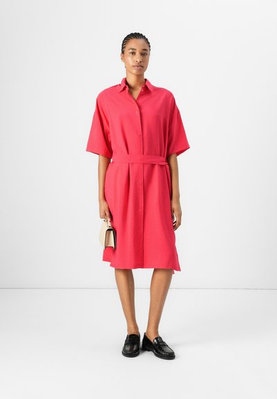 Robe chemise rose avec une ceinture, dotée de manches courtes, d'un devant à boutons et d'une coupe loose. Des mocassins noirs et un petit sac beige complètent le look.