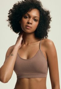 Brązowy bawełniany biustonosz braletka z cienkimi ramiączkami, o bezszwowym designie i gładkiej fakturze. Modelka pozuje z ręką na szyi.