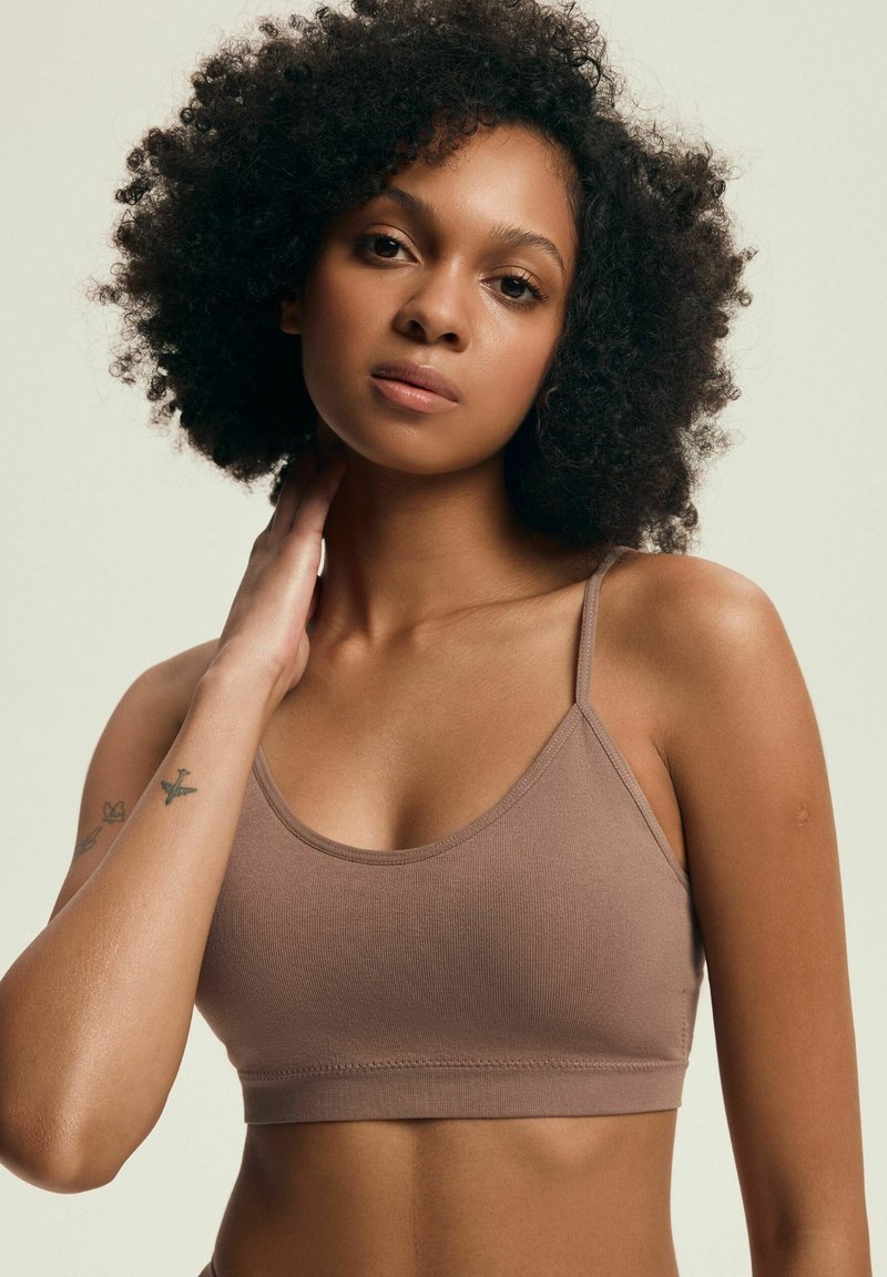 Brązowy bawełniany biustonosz braletka z cienkimi ramiączkami, o bezszwowym designie i gładkiej fakturze. Modelka pozuje z ręką na szyi.