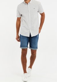 Camisa de botones de manga corta, de color gris claro, con una tela texturizada y un bolsillo frontal, combinada con pantalones cortos de denim azul enrollados y zapatillas blancas.