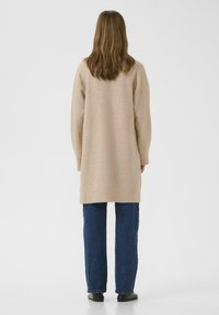 Moters su ilgais rudais plaukais, vilkinčios smėlio spalvos ilgą kardiganą, mėlynas džinsus ir juodas batus, stovinčios atgręžtos nuo objektyvo prieš paprastą fono.