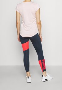 Top rosa chiaro a maniche corte abbinato a leggings blu navy con una vivace striscia arancione e il logo "PUMA" sulla gamba inferiore, con scarpe da ginnastica bianche.
