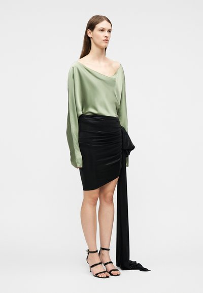 Γυναίκα που φοράει μια αέρινη μπλούζα από σατέν σε απαλό πράσινο χρώμα με έναν ώμο ακάλυπτο, μια μαύρη μίνι φούστα με πιέτες και μακριά πλευρική επίστρωση, και μαύρα πέδιλα με λουράκια.