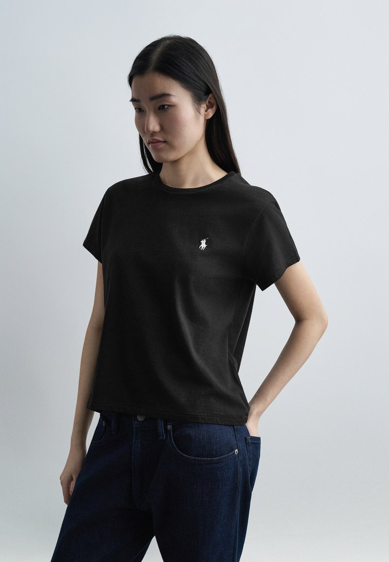 Νεαρή γυναίκα που φοράει μαύρο μπλουζάκι με κοντά μανίκια και μικρό λευκό λογότυπο στο στήθος και σκούρο μπλε τζιν, στέκεται μπροστά σε ανοιχτό γκρι φόντο.