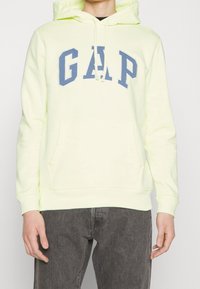 Lichtgele hoodie met een blauwe "GAP" logo op de borst, voorzien van een voorzak en een trekkoord in de capuchon. Zachte, iets gestructureerde stof.