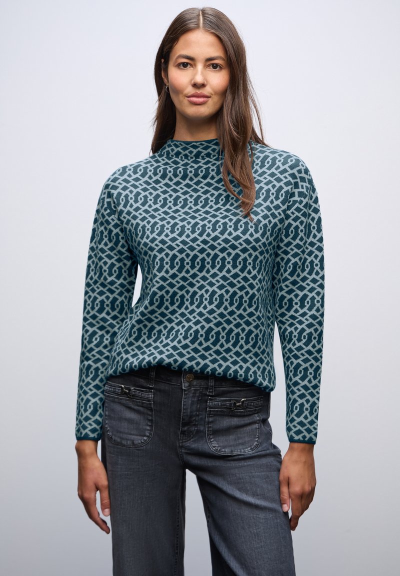 Top in maglina turchese con scollatura alta e motivo geometrico. Abbinato a jeans grigio scuro con dettagli con zip. Morbido al tatto.