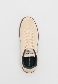 Baskets en daim beige à lacets avec semelle foncée, marquage Lacoste sur la languette et à l’intérieur de la semelle, et accent vert sur le côté.