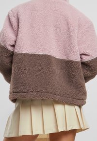 Chaqueta de forro polar con la parte superior rosa y la inferior marrón, que presenta una superficie texturizada y un detalle de piping blanco a lo largo de la costura.