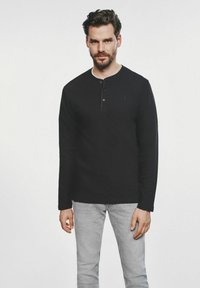 Czarna koszula typu henley z długim rękawem, o teksturowanej tkaninie, z plisą guzikową i małym haftowanym logo na piersi, zestawiona z jasnoszarymi dżinsami.