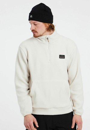 White fleece pullover with a half zip, front pouch pocket, and a black brand patch. Features ribbed cuffs and a textured, soft surface.