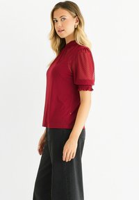 Red short-sleeve blouse with a dotted pattern, featuring a high neckline and ruffled sleeves, paired with wide-leg black pants.
