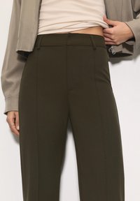 Mørkegrønne skræddersyede bukser med lige snit, glat front og ingen synlige lommer. Stoffet virker glat og struktureret.