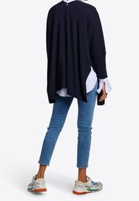 Námořnicky modrý pletený pončo s kulatým tvarem, opatřený zapínáním na knoflík na zádech. Nošený přes bílou pruhovanou košili a kombinovaný s modrými džínami a teniskami.