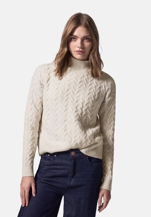 Pull en maille torsadée crème avec col montant et poignets côtelés, associé à un jean en denim foncé. Le pull présente un design texturé et épais.