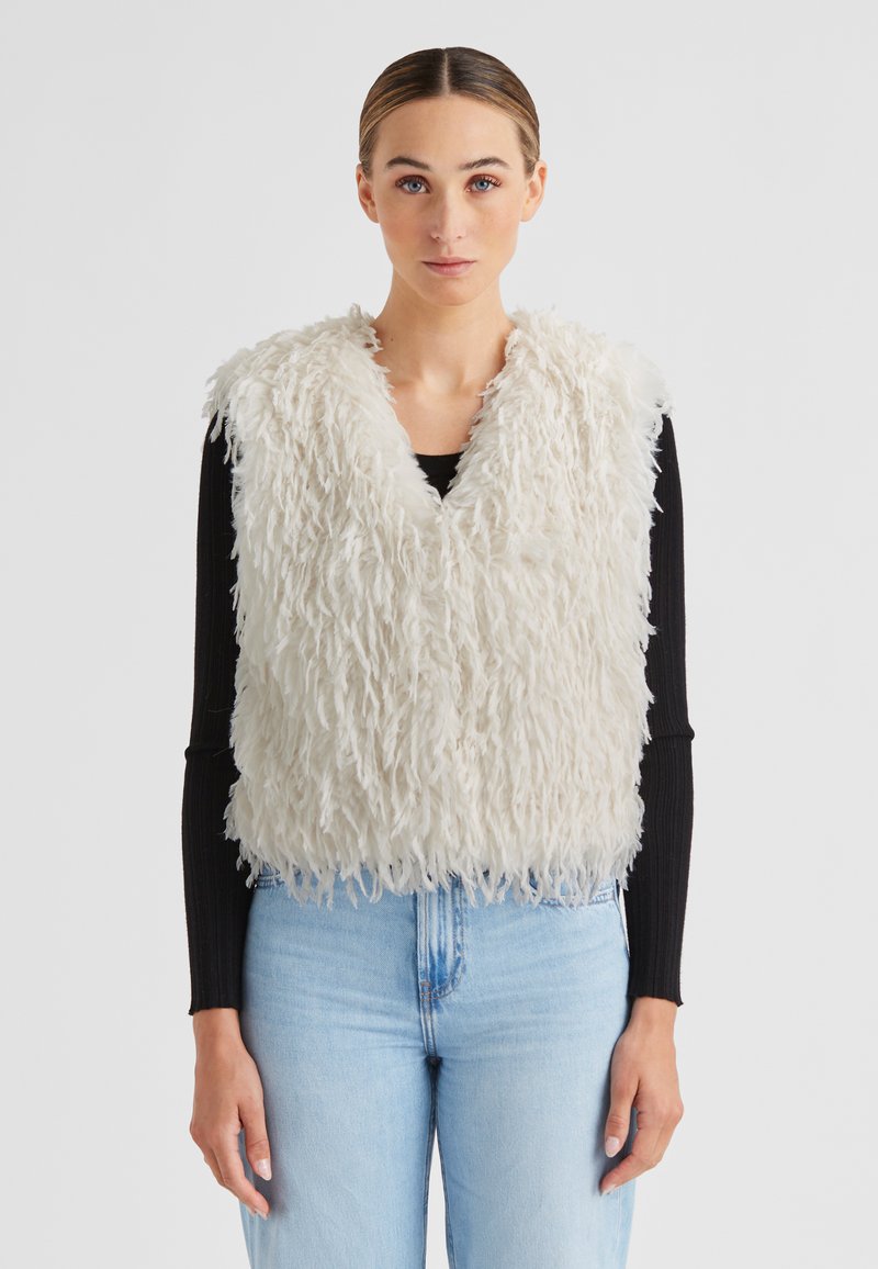 Kunstpelz-Weste in Off-White mit V-Ausschnitt. Strukturierte, flauschige Oberfläche mit einem breiten Saum. Über einem schwarzen Langarmoberteil und blauen Jeans getragen.