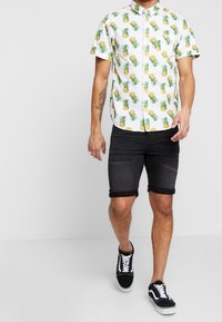 Camisa de botones de manga corta con fondo blanco y patrón de piñas amarillo-verdosas; combinada con pantalones cortos de mezclilla negros arremangados y zapatillas negras.
