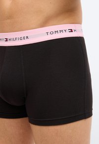 Κοντινό πλάνο ενός ατόμου που φοράει μαύρα μπόξερ με ροζ μέση που φέρει το λογότυπο Tommy Hilfiger.