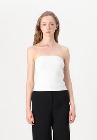Bijela rebrasta top bez naramenica, slim fit, s glatkom teksturom. Uparen s crnim hlačama širokih nogavica, stvarajući kontrastni outfit.