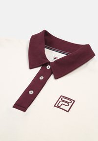 Paita polo vaaleassa kermaassa, jossa on burgundinvärinen kaulus ja viisi nappia. Vasemmassa rinnassa on pieni brodeerattu logo. Pehmeä tekstiili, klassinen muotoilu.