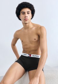 Μαύρα βαμβακερά μποξεράκια με λευκή ελαστική ζώνη μέσης που φέρει το λογότυπο «TOMMY HILFIGER», παρουσιάζονται σε αντρικό μοντέλο με φόντο ανοιχτόχρωμο.