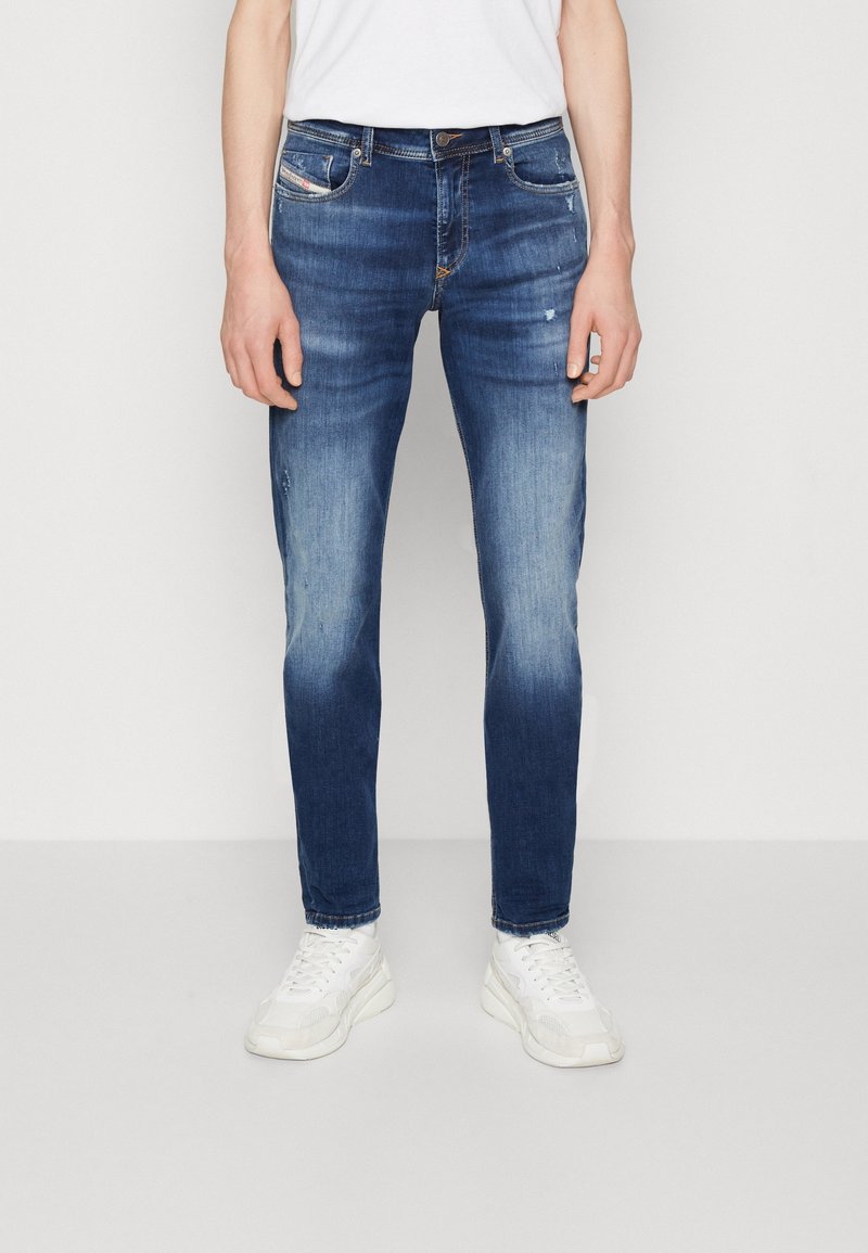 Jeans de mezclilla azul ajustados con detalles desgastados y deslavados, diseño de cinco bolsillos y cintura de altura media, combinados con zapatillas blancas.