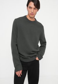 Ciemnoszary sweter z okrągłym dekoltem i prążkowanymi mankietami, wykonany z materiału o fakturowanym splocie, w zestawie z czarnymi dżinsami.