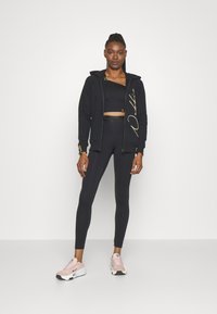 Fekete zipzáros kapucnis pulóver arany logóval, fekete crop top-pal és magas derekú leggings-szel párosítva. Világos rózsaszín sportcipők fehér részletekkel.