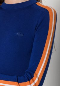 Blå långärmad skjorta i stickad tyg med orangea och vita ränder längs ärmarna. Litet, broderat logo på bröstet.