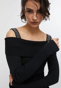 Črna rebrasta majica z dolgimi rokavi, izrez na ramenih, okrašena s srebrnimi zakovicami na naramnicah in teksturirano tkanino. Tesno prilegajoč kroj z minimalnimi detajli.