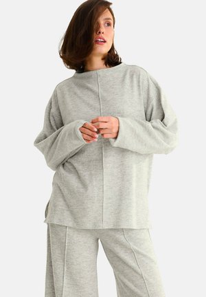 Sweatshirt oversized gris clair, avec des manches longues, une texture douce, un col large et des détails de couture diagonale, porté avec un pantalon assorti.