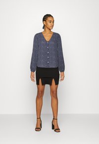Blusa azul-marinho com bolinhas brancas, decote em V e detalhes em botão, combinada com uma saia mini preta com uma fenda frontal, completada com saltos pretos.