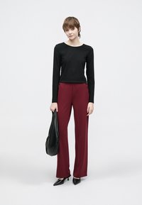 Μαύρο πουκάμισο με μακριά μανίκια, μπουρντό παστέλ παντελόνι με διακοσμητικά κουμπιά στο πλάι, μαύρη τσάντα χειρός και μυτερά μαύρα τακούνια πάνω σε ανοιχτό φόντο.