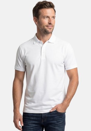 Mann mit kurzem braunem Haar, der ein weißes kurzärmeliges Poloshirt und eine dunkle Jeans trägt, mit einer Hand in der Tasche stehend.