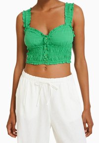 Zielony krótki top z falbaniastymi ramiączkami, teksturowany materiał, z guzikiem z przodu, połączony z białymi spodniami ze sznurkiem.