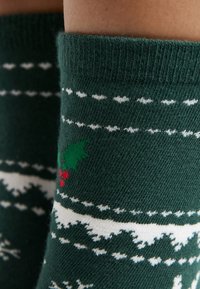 Zielone wełniane skarpetki z białym i czerwonym wzorem świątecznym, zawierające liście ostrokrzewu i jagody. Miękka faktura z prążkowanym mankietem.