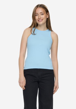 Joven sonriente, vestida con una camiseta sin mangas azul claro y pantalones negros, de pie frente a un fondo blanco liso.