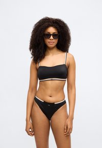 Melns bikini komplekts ar bandeau tipus augšdaļu, plānām lencītēm un dekoratīvām detaļām, kā arī zemu jostas līniju biksītēm ar līdzīgām detaļām.