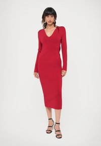 Rochie midi tricotată roșie, cu mâneci lungi și decolteu în V, silueta ajustată, textură netedă și sandale negre cu bretele la gleznă.