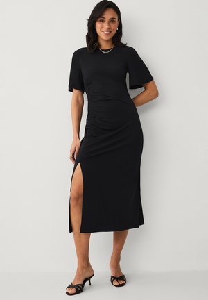 Kvinde iført en sort midi-kjole med korte ærmer, sidesamlinger, høj slids på låret, sorte hælede sandaler og minimal smykker.