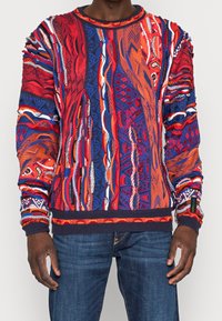 Man in blauwe jeans en een levendige, kleurrijke gebreide trui met abstracte patronen in rood, oranje, blauw en wit.