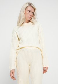 Pulover crem tricotat cu model împletit și pantaloni asortați dintr-un tricot moale, texturat. Ținuta are un croi lejer și manșete tricotate, creând un aspect confortabil.