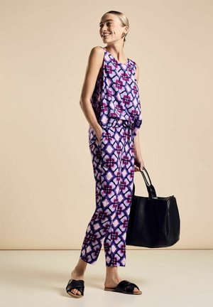 Completo due pezzi viola con motivo, composto da un top morbido e pantaloni a sigaretta; presenta design geometrici e tasche; abbinato a una borsa nera e sandali.